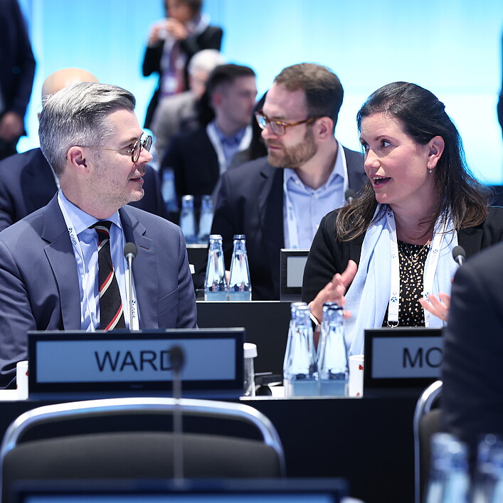 Mehrere Personen sitzen an Tischen in einem modern beleuchteten Konferenzsaal. Im Vordergrund unterhalten sich zwei Teilnehmende miteinander. Auf den Tischen stehen Wasserflaschen, Mikrofone und Namensschilder. Im Hintergrund sind weitere Teilnehmende zu sehen.