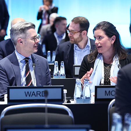 Mehrere Personen sitzen an Tischen in einem modern beleuchteten Konferenzsaal. Im Vordergrund unterhalten sich zwei Teilnehmende miteinander. Auf den Tischen stehen Wasserflaschen, Mikrofone und Namensschilder. Im Hintergrund sind weitere Teilnehmende zu sehen.