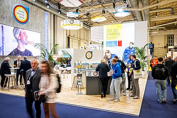  Messestand zum Thema „Sanitär, Heizung, Klima“ mit großem Bildschirm und dem Hashtag „#WICHTIGESTUER“. Ein Schild zeigt „Zeit zu starten“, Personen sind im Gespräch oder laufen vorbei.