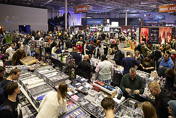 Große Messehalle mit vielen Menschen, die an dicht gefüllten Verkaufsständen vorbeigehen. Auf den Tischen liegen zahlreiche Spiele, Sammelkarten, Figuren und Merchandise-Artikel. Im Hintergrund sind weitere Bereiche der Veranstaltung mit Schildern wie ‚Retro Area‘, ‚Cybedrome‘ und ‚Indie Area‘ zu sehen. Die Halle ist hell ausgeleuchtet und stark besucht.