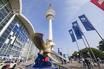 View of the Hamburg exhibition grounds with the Heinrich Hertz Tower in the center. A large ship propeller sculpture is displayed in the foreground, while several blue trade fair flags are flying on the right. Visitors are walking near the Hamburg Messe & Congress entrance under a clear blue sky.