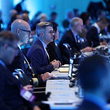 Reihe von Teilnehmenden bei einer Konferenz oder Sitzung, die an langen Tischen mit Mikrofonen und Laptops sitzen und aufmerksam nach vorne ausgerichtet sind; der Raum ist in kühles, blaues Licht getaucht, im Hintergrund sind weitere unscharfe Personen und Technik zu erkennen.