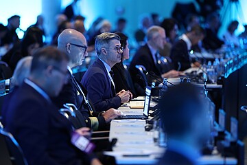 Teilnehmerinnen und Teilnehmer sitzen in mehreren Reihen an langen Tischen in einem Konferenzsaal. Vor ihnen stehen Laptops, Mikrofone und Unterlagen. Die Beleuchtung ist kühl blau, und die Atmosphäre wirkt konzentriert und professionell.