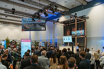 Besucherinnen und Besucher verfolgen eine Präsentation auf einer Logistik-Fachveranstaltung vor einer Bühnen- und Ausstellungsfläche.