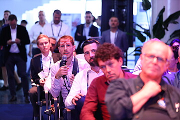 A person is seated in the audience at an event, holding a microphone while asking a question or making a comment. The setting appears to be a busy conference or networking space with several attendees seated and standing in the background, listening attentively.