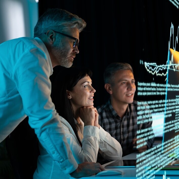 Three professionals collaboratively examine a technical device containing visible circuit boards in a modern engineering or laboratory environment. Computer monitors displaying technical information are positioned on the desk, while a task lamp illuminates the prototype. Additional technical equipment and a robotic arm can be seen in the background.