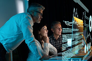 Three professionals collaboratively examine a technical device containing visible circuit boards in a modern engineering or laboratory environment. Computer monitors displaying technical information are positioned on the desk, while a task lamp illuminates the prototype. Additional technical equipment and a robotic arm can be seen in the background.