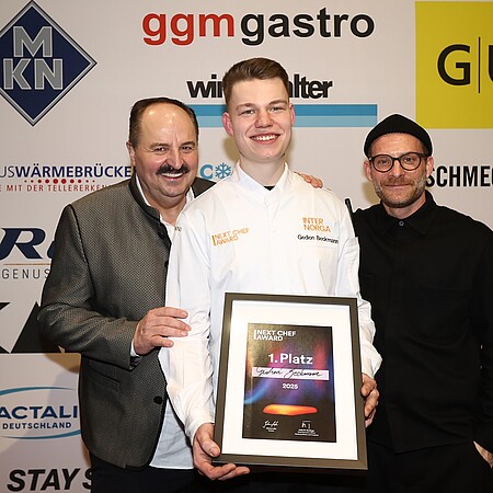 Der Gewinner des Next Chef Awards 2025, Gedion Beckmann, hält stolz seine Siegerurkunde, während Sternekoch Johann Lafer (links) und Moderator Sebastian E. Merget (rechts) ihn gratulierend flankieren.