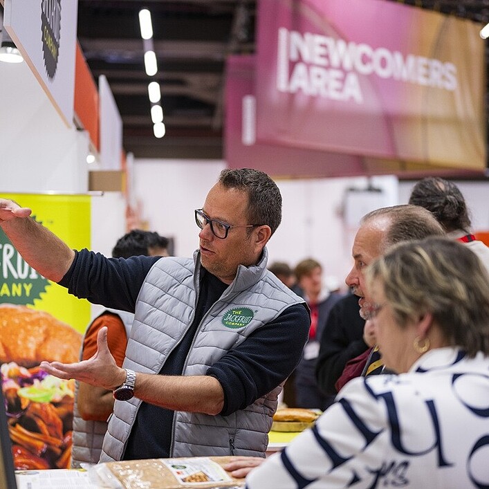 Eine Person auf einer Messe erklärt einer kleinen Gruppe von Besucherinnen und Besuchern ausführlich ein Produkt an einem Messestand. Im Hintergrund sind bunte Banner und Schilder zu sehen, darunter ein Bereich mit der Aufschrift Newcomers Area. Auf dem Tisch liegen verschiedene Lebensmittelproben und Informationsmaterialien.