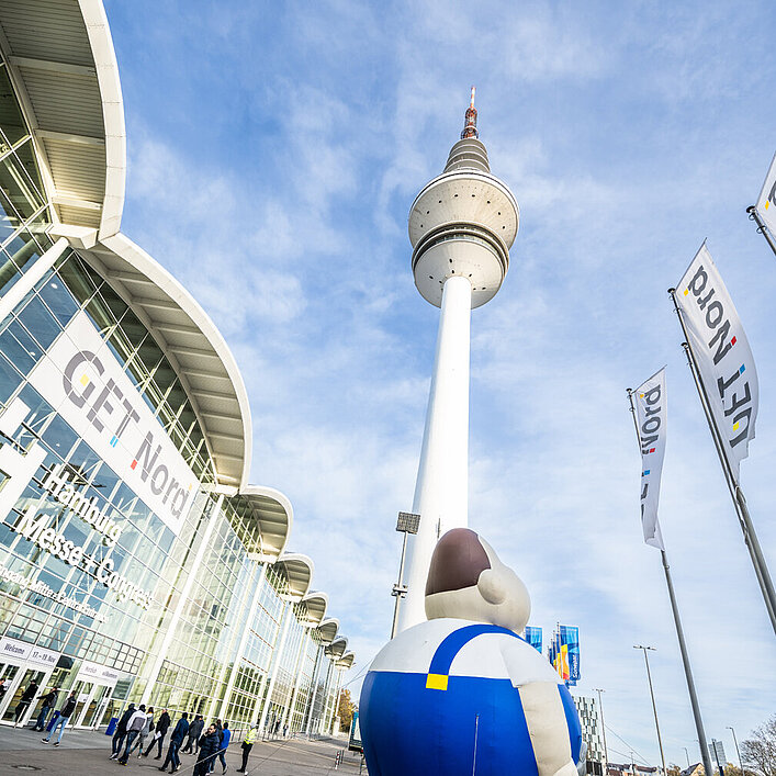 Das Messegebäude ist zu sehen, der Hamburger Fernsehturm und drei Get Nord Flaggen. Personen laufen draußen umher und es ist eine große aufgeblasene Figur zu sehen. 