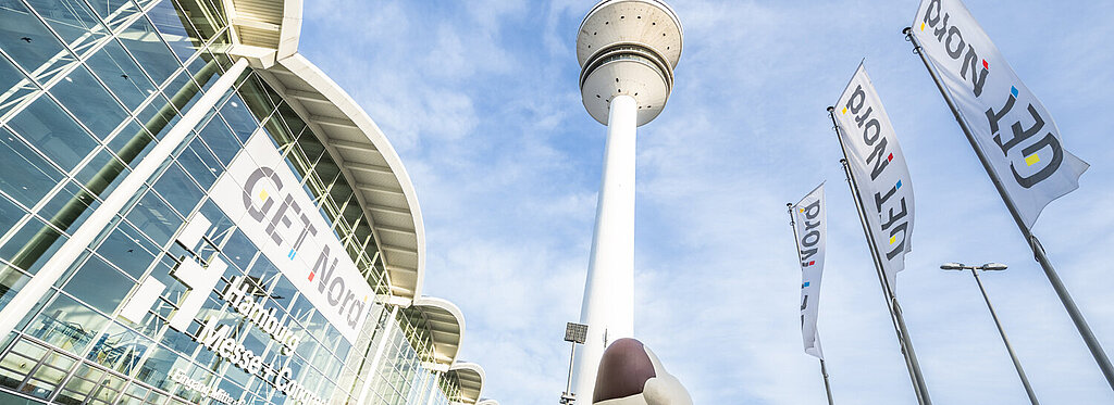 Das Messegebäude ist zu sehen, der Hamburger Fernsehturm und drei Get Nord Flaggen. Personen laufen draußen umher und es ist eine große aufgeblasene Figur zu sehen. 