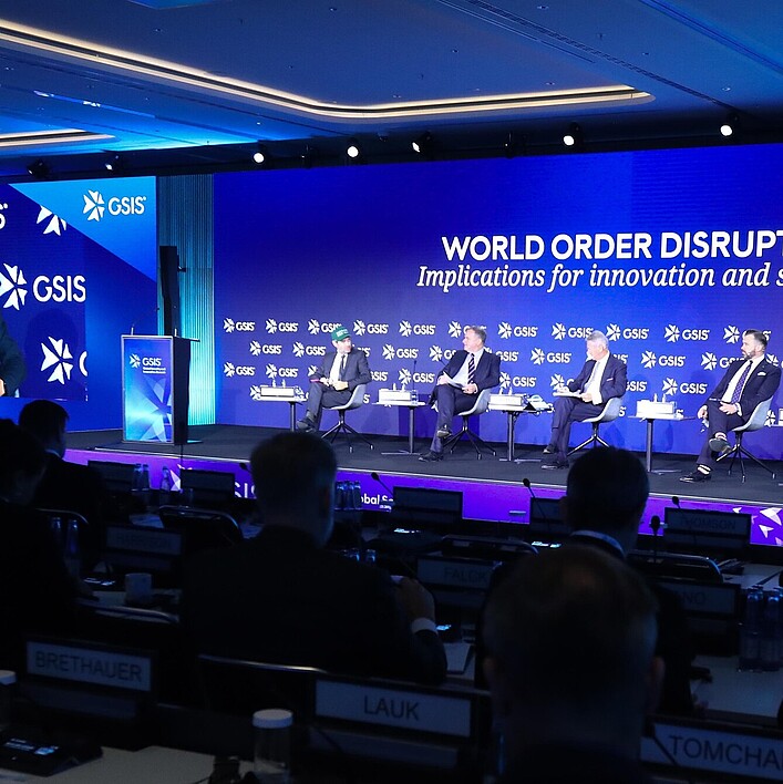 Panel discussion at an international GSIS conference: several people are seated on a stage in front of a large blue screen displaying the text ‘World Order Disrupted: Implications for innovation and security.’ On the left, a speaker stands at a podium, while their image is shown on a large projection screen. In the foreground, audience members are visible in a conference hall