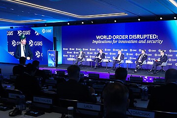 Panel discussion at an international GSIS conference: several people are seated on a stage in front of a large blue screen displaying the text ‘World Order Disrupted: Implications for innovation and security.’ On the left, a speaker stands at a podium, while their image is shown on a large projection screen. In the foreground, audience members are visible in a conference hall