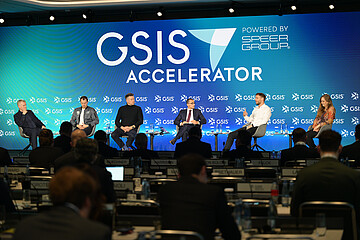 Podiumsdiskussion auf der GSIS Accelerator Conference vor einem großen, blau beleuchteten Bühnenhintergrund mit GSIS-Logo. Mehrere Teilnehmende sitzen auf der Bühne und diskutieren vor einem Publikum.