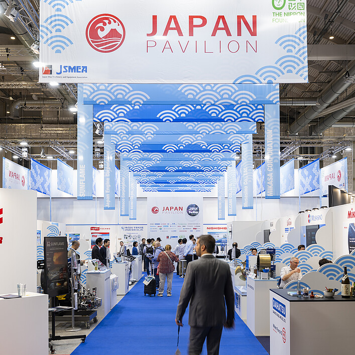View through the Japan Pavilion in an exhibition hall, showing a central blue carpet leading past maritime industry booths. Overhead banners with wave patterns and the text “Japan Pavilion” hang from the ceiling.