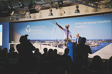 Person spricht an einem Rednerpult vor Publikum auf einer Bühne der WindEnergy Hamburg. Im Hintergrund ist eine großformatige Veranstaltungswand mit WindEnergy-Hamburg-Logo und Hamburg-Panorama zu sehen.