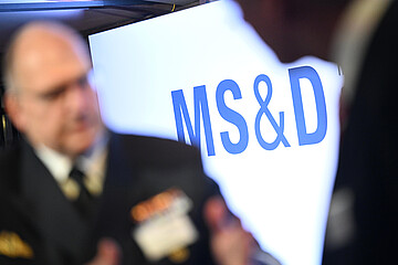Unscharfes Foto einer Veranstaltungsszene: Im Vordergrund steht ein deutlich sichtbares blaues „MS&D“-Logo auf einer hellen Fläche, während im Hintergrund eine Person in dunkler Kleidung mit Abzeichen stark verschwommen zu sehen ist.