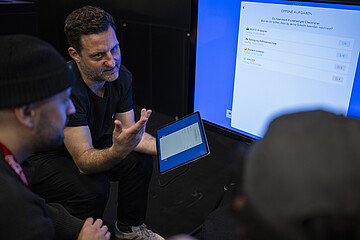 Drei Personen besprechen die digitale Verwaltung offener Aufgaben. Auf einem großen Bildschirm und einem Tablet ist eine Checkliste mit noch zu erledigenden Aufgaben zu sehen.