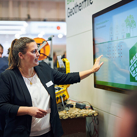 Eine Frau präsentiert auf einer Messe eine große digitale Anzeige zum Thema Geothermie. Sie zeigt auf ein Diagramm, während mehrere Personen aufmerksam zuhören.