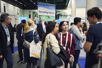 Messebesuchende im Gespräch an einem Informationsstand in einer gut besuchten Messehalle.