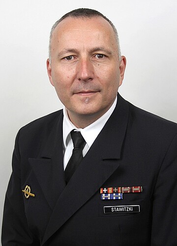 Person in dunkler Uniform vor hellem Hintergrund, mit mehreren Abzeichen und einem Namensschild, das den Text ‚STAWITZKI‘ zeigt.