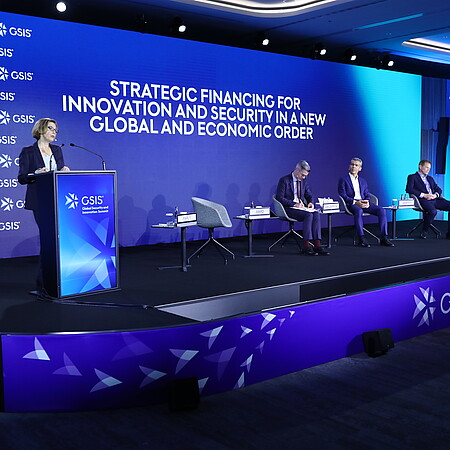Conference stage at the GSIS (Global Security and Innovation Summit) in Hamburg. A speaker stands at a podium in front of a large blue LED screen displaying the title “Strategic Financing for Innovation and Security in a New Global and Economic Order.” Several participants are seated on stage as part of a panel discussion. A large screen on the right shows the speaker. The event takes place in a modern, professionally lit conference venue.