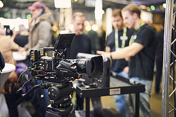 Professional video camera at an exhibition stand during a trade fair event, with attendees interacting and networking in the background.
