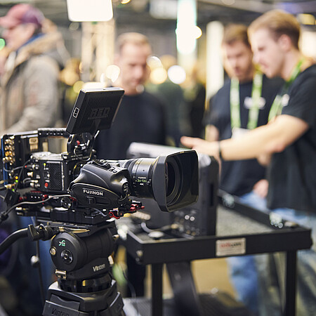 Professional video camera at an exhibition stand during a trade fair event, with attendees interacting and networking in the background.