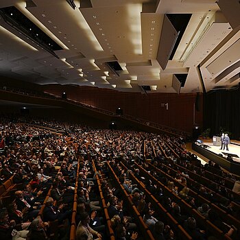 A fully packed auditorium with a modern ceiling. Two people are standing on a stage, and their images are projected onto a large screen in the background.