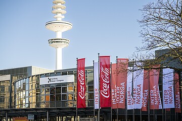 Außenansicht des Hamburg Messe und Congress Gebäudes bei klarem Himmel. Vor dem Eingang stehen mehrere rote und weiße Fahnen mit Coca-Cola- und INTERNORGA‑Branding. Im Hintergrund ragt der markante Heinrich-Hertz-Turm in den Himmel.