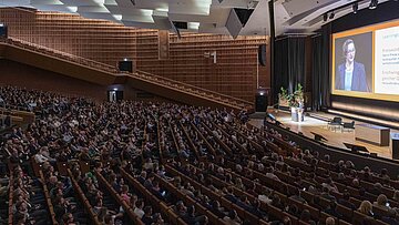 „Eine voll besetzte Konferenzhalle mit moderner Holzarchitektur. Auf der Bühne spricht eine Person am Rednerpult, während ihre Präsentation auf einer großen Leinwand im Hintergrund projiziert wird.“