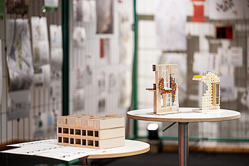 Architekturmodelle und Materialstudien auf zwei runden Tischen vor einer Ausstellungswand mit Entwürfen.