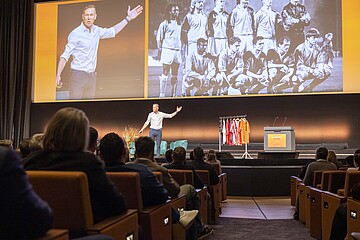 Person auf einer Bühne vor Publikum während einer Konferenzpräsentation. Im Hintergrund zeigt eine große Leinwand ein historisches Mannschaftsfoto sowie eine vergrößerte Aufnahme der präsentierenden Person. Rechts neben der Bühne stehen mehrere Trikots auf einem Kleiderständer und ein Rednerpult.