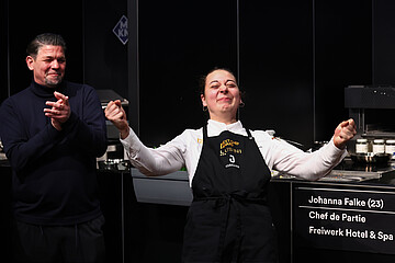 Next Chef Award: Eine Teilnehmerin des Next Chef Award steht in Kochkleidung auf der Bühne, während eine weitere Person daneben applaudiert.