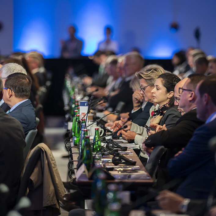 Participants are seated at long tables during a conference, with microphones, notes, and water bottles in front of them, while additional attendees listen in the background.
