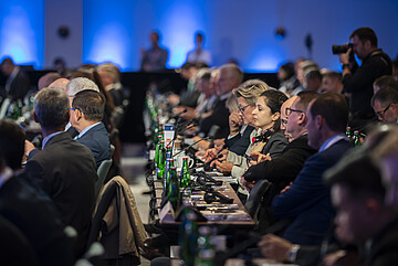 Participants are seated at long tables during a conference, with microphones, notes, and water bottles in front of them, while additional attendees listen in the background.