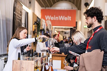 Eine Person schenkt an einem Messestand ein Getränk in ein Glas ein, während mehrere Besucherinnen und Besucher davor stehen. Im Hintergrund hängt ein großes Schild mit der Aufschrift „WeinWelt“.