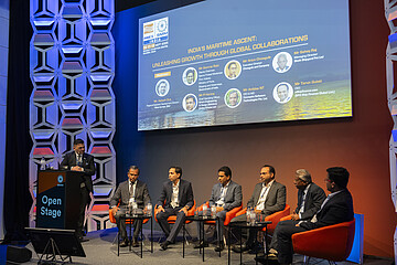 Panel discussion on a conference stage. One person stands at a lectern labeled “Open Stage” while six other participants are seated in orange chairs. A large screen in the background displays information about a maritime industry event in India. The stage is lit in blue and orange tones and framed by decorative wall panels.