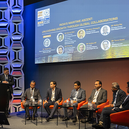 Panel discussion on a conference stage. One person stands at a lectern labeled “Open Stage” while six other participants are seated in orange chairs. A large screen in the background displays information about a maritime industry event in India. The stage is lit in blue and orange tones and framed by decorative wall panels.