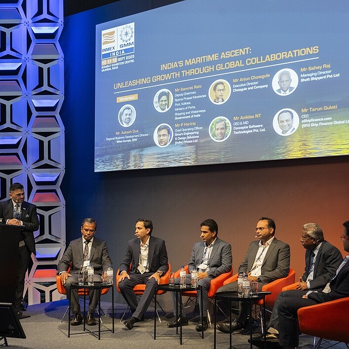 Eine Podiumsdiskussion auf einer Bühne mit sieben Personen, von denen eine am Rednerpult steht und sechs in einer Reihe auf orangefarbenen Sesseln sitzen. Hinter ihnen zeigt eine große Leinwand den Titel der Veranstaltung zum Thema maritime Entwicklung und internationale Zusammenarbeit. Die Bühne ist modern ausgeleuchtet und hat eine geometrische Wandgestaltung. Das Publikum ist nicht sichtbar.