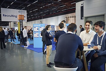 Menschen befinden sich auf einer Messefläche im Recruiting‑Bereich; einige stehen vor einer Jobwall, andere sitzen an einem Stehtisch und unterhalten sich.