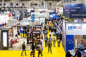 Innenaufnahme einer Messehalle mit zahlreichen Ausstellern und Besuchern. Die Halle ist hell beleuchtet, mit gelbem Boden. Zu sehen sind zahlreiche Stände.