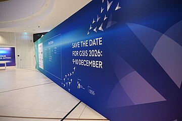 Große blaue Messewand in einem modernen Innenraum mit dem Schriftzug „Save the Date for GSIS 2026: 9–10 December“ sowie grafischen Dreiecksformen.